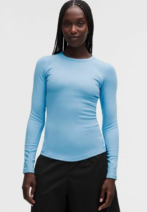 Frau mit langem geflochtenem Haar, die ein eng anliegendes hellblaues Langarmshirt und schwarze Hose trägt, steht vor einem schlichten Hintergrund.