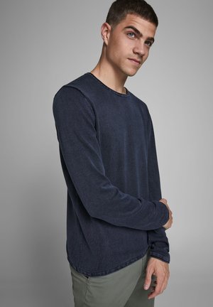 Camisa de manga larga azul marino en un tejido texturizado con cuello redondo y detalles cosidos. Llevada con pantalones verde claro.