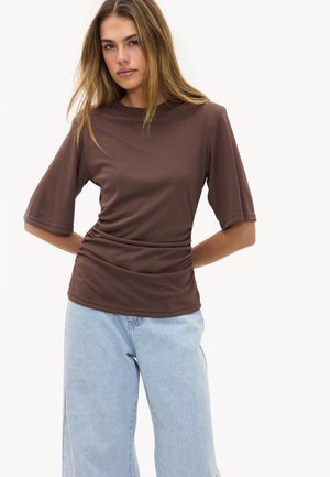 Young woman with long hair wearing a brown ruched short-sleeve top and light blue wide-leg jeans, standing with hands behind back.