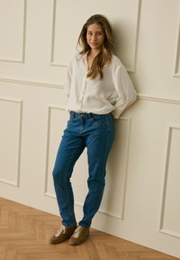 Femeie cu păr lung și maro, purtând o bluză albă, blugi albaștri și adidași maro, sprijinită lejer de un perete crem cu panouri.