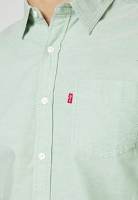 Chemise verte claire à boutons avec une poche poitrine et une étiquette en tissu rouge Levi's visible sur le bord de la poche.