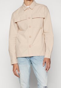 Chaqueta beige de botón con cuello, dos bolsillos frontales y mangas largas. Tejido texturizado con detalle de ligeras estrías.