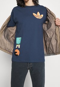 Námořnické bavlněné tričko s oranžovým maskáčovým logem na hrudi a dvěma barevnými grafickými nášivkami na přední straně, v uvolněném střihu.