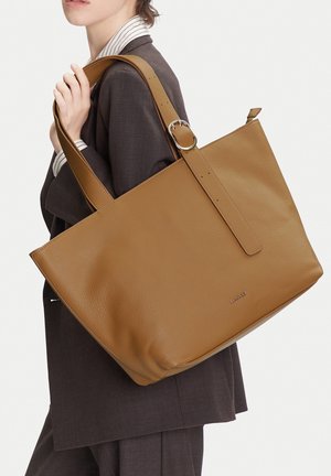 Persona con blazer oscuro lleva un gran bolso tote de cuero marrón con correa de hebilla sobre el hombro, con un fondo liso.