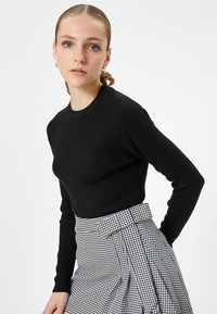 Czarny, prążkowany sweter z długim rękawem w połączeniu z spódnicą w czarno-białą kratę w stylu hounds-tooth, z bocznymi plisami i akcentem w postaci kokardy.