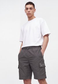 Jeune homme portant un t-shirt blanc uni à manches courtes et un short cargo gris, les mains dans les poches, debout devant un fond blanc.