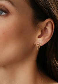 Guldtonade örhängen med ett båtdesign med böjda ändar, placerade mot en neutral hudton, vilket framhäver smyckets släta textur.