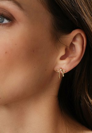 Orecchini in tono oro con un design a fiocco dalle estremità curve, posizionati su una tonalità di pelle neutra, evidenziando la texture elegante dei gioielli.