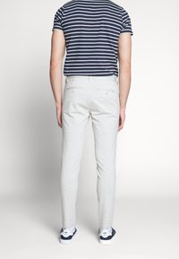 Light grey trousers with a slim fit, featuring two back pockets with button closures. Worn with a navy and white striped t-shirt and sneakers.