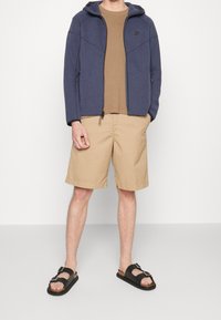 Marin zip-up hoodie med en texturerad yta, brun åtsittande t-shirt, beige shorts och svarta sandaler med bred rem och spänne.