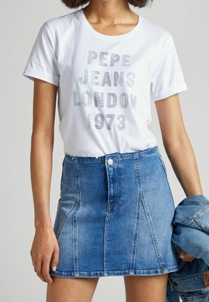 Frau trägt ein weißes T-Shirt mit dem Aufdruck "Pepe Jeans London 1973" und einen blauen Jeansrock, hält eine gefaltete Jeansjacke in einer Hand.