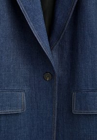 Tumman sininen farkkutakki, jossa on lovelline kaulus, yksi musta nappi ja kaksi etutaskua. Kontrastivärinen ommel tuo muotoa ja rakennetta.