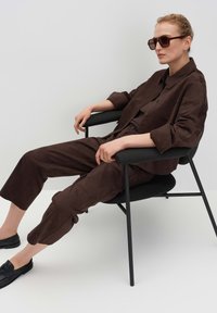 Braunes Wildleder-Zweiteiler-Set mit einem Knopfhemd und kurzen Hosen. Modell sitzt auf einem schwarzen Stuhl, trägt dunkle Sonnenbrillen und Slipper.
