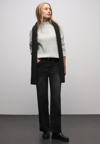 Maglione grigio chiaro a coste con motivo a diamante, abbinato a jeans neri a vita alta e una lunga sciarpa nera. Completa il look un paio di stivaletti neri alla caviglia.