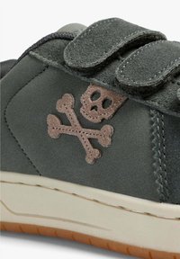 Zapatillas de ante verde con correas de Velcro. Presenta un logo de calavera y huesos cruzados en color beige en el lateral, una entresuela blanca y una suela de goma marrón.