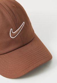 Brun stoff baseballcaps med hvit brodert Nike swoosh-logo på fronten, detaljert søm på buet skygge.