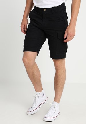 Homme portant un short cargo noir et des baskets montantes blanches, debout sur un fond uni.