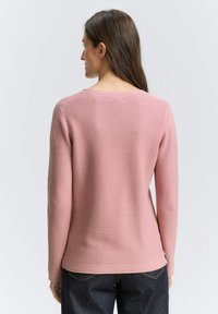 Vrouw met lang bruin haar, gekleed in een lichte, geribbelde top met lange mouwen in zachtroze en donkere jeans, met haar rug naar de camera gekeerd tegen een lichte achtergrond.