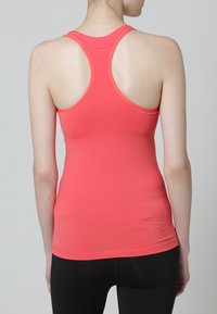 Nainen, joka on pukeutunut korallinväriseen urheilulliseen hihattomaan paitaansa ja mustiin legginsseihin, näkyy selkäpuolelta yksinkertaista harmaata taustaa vasten.