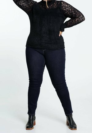Femme portant un haut noir en dentelle à manches longues, un jean skinny foncé et des bottines noires à talons en bois, debout avec une main sur la hanche.