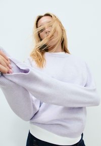 Blonde woman with hair partially covering face, wearing a light purple sweater and adjusting the sleeve against a plain white background.