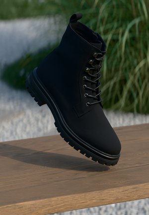 Musta nilkkaboots, joka on valmistettu sileästä materiaalista. Ominaisuuksiin kuuluvat nauhakiinnitys, pyöreä kärki ja kumipohja, jossa on pitävä kuvio.