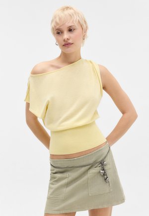 Lühikeste blondide juustega naine, kes kannab kahvatukollast õlavaba topi ja beeži seelikut hõbedase südamekujundusega vööga, seisab käed selja taga.