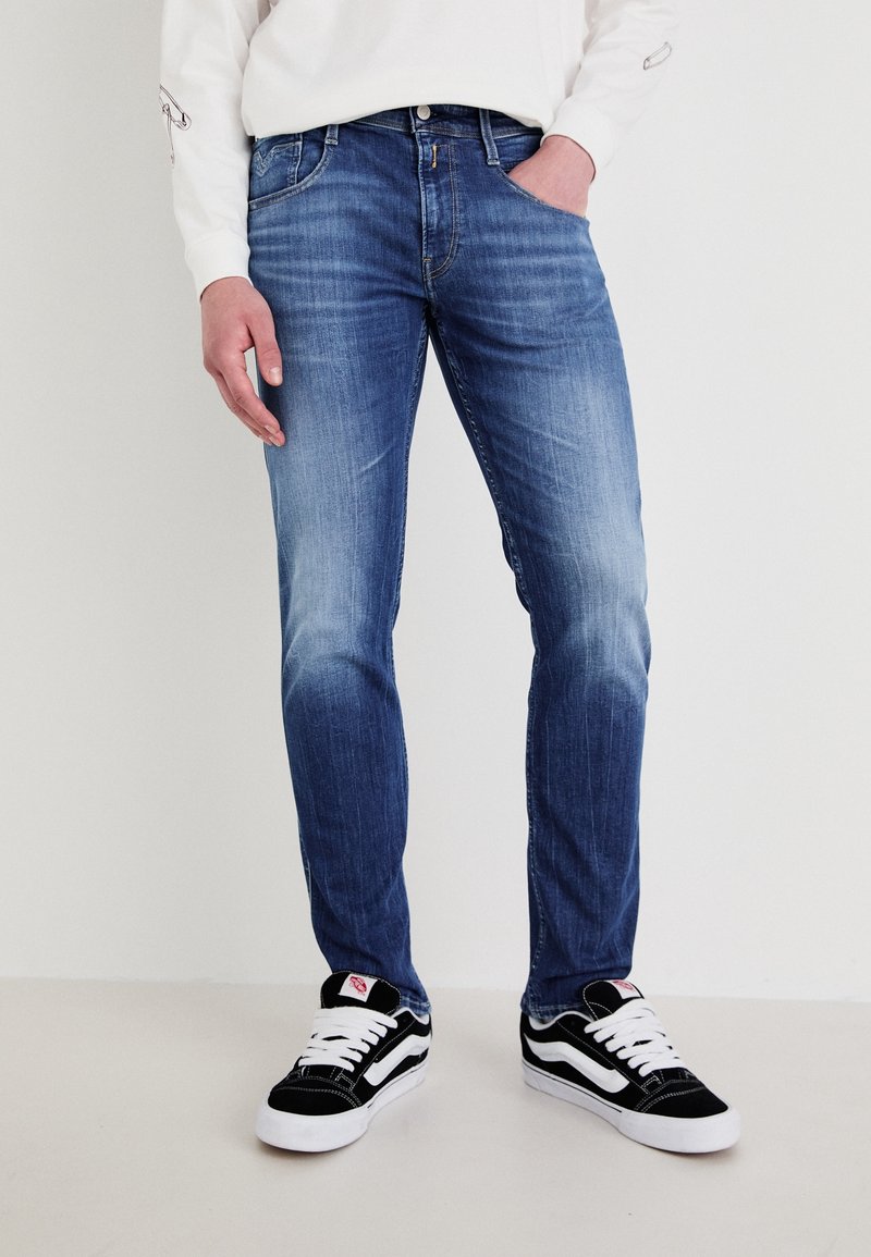 Personne portant un jean slim délavé bleu et des baskets noires et blanches, debout avec une main dans la poche devant un fond uni.
