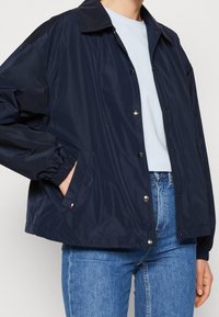 Granatowa nylonowa kurtka z zapięciem na zatrzaski, elastycznymi mankietami i bocznymi kieszeniami, charakteryzująca się casualowym stylem i gładką teksturą.