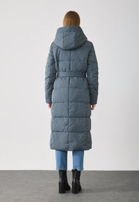 Γυναίκα π�ου φοράει ένα μακρύ, ζωνάκι, επενδυμένο γκρίζο παλτό με κουκούλα, μπλε κοντά τζιν και μαύρες ψηλές μπότες μέχρι τον αστράγαλο, στέκεται σε γκρι πάτωμα.