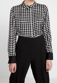 Bluse med sort-hvid houndstooth mønster, krave og knaplukning, med lange ærmer og sorte mansjetter.