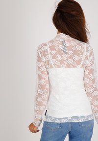 Witte kanten top met bloemmotieven, lange mouwen en een transparante achterkant. Beschikt over een gladde witte onderlaag en een rechte zoom. Jeans afgebeeld.