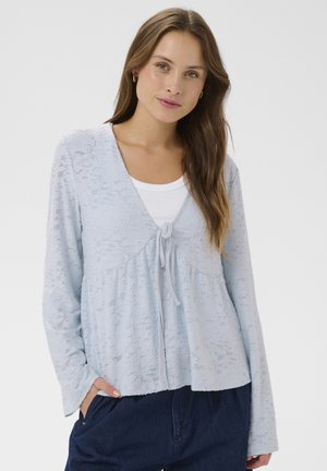 Ženska z dolgimi rjavimi lasmi, oblečena v svetlomodri čipkasti kardigan z rožnim vzorcem preko belega zgornjega dela in temno modrih hlačah, z rokami v žepih, na enobarvnem ozadju.