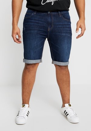 Mężczyzna ubrany w ciemnoniebieskie, podwinięte szorty dżinsowe, czarny t-shirt oraz białe trampki z czarnymi paskami, stojący na jednolitym tle.