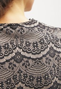 Svart spetstyg har intrikata blommönster och vågiga kanter, lager över en nude bakgrund, vilket skapar en delikat struktur.