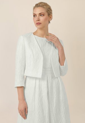 Frau mit blondem Haar trägt eine strukturierte weiße, bauchfreie Jacke über einem passenden Kleid, steht vor beigefarbenem Hintergrund und blickt nach links.