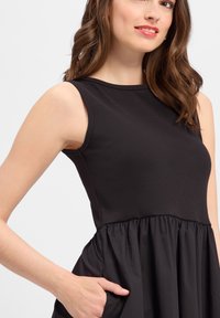 Schwarzes Kleid mit einem taillierten, gerippten Oberteil und einem ausgestellten, rüschenden Rock. Enthält Seitentaschen und einen Rundhalsausschnitt. Satinähnliche Textur.