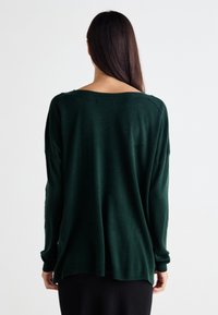 Pulover supradimensionat de culoare verde închis, cu un guler larg, mâneci lungi și o croială lejeră. Textură moale cu accente ribbate la manșete și la tiv.