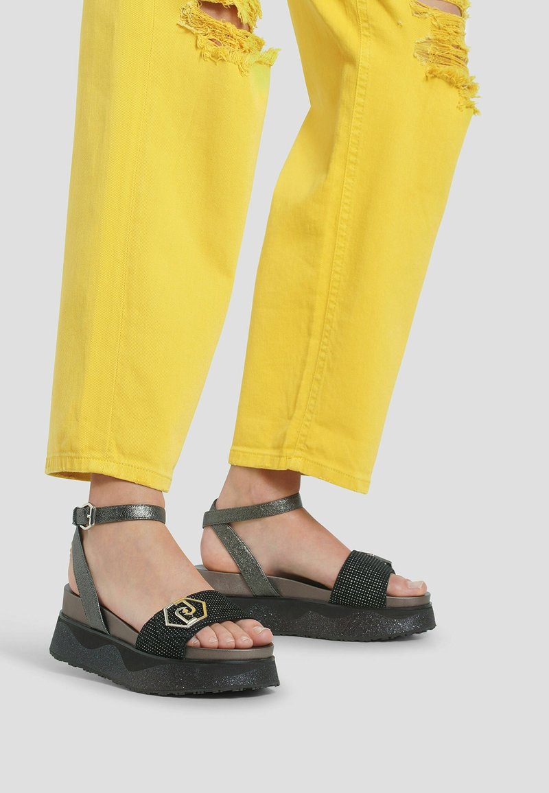 Sandales noires et argentées avec une bride texturée et un détail logo. Semelle compensée légèrement scintillante, portées avec un jean jaune déchiré.