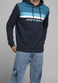 Laivastonsininen huppari, jossa on teksturoitu sininen yläpaneeli, jossa on valkoisia ja vaaleansinisia raitoja sekä "JACK & JONES" -teksti painettuna paksulla fontilla.
