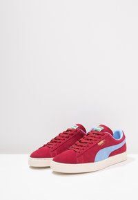 Röda mockaskor med ljusblå accenter, vit gummisula och platta snören. Har logotyp på tungan och sidan. Avslappnad design.