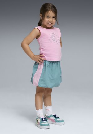 Młoda dziewczynka stoi uśmiechnięta z rękami na biodrach, ubrana w różowy bezrękawnik, turkusową spódnicę z różowym paskiem, białe skarpetki i wielokolorowe trampki.