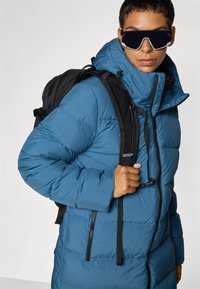 Teal puffer coat with a high collar, zip closure, and black backpack straps. Features quilted texture and a secure front pocket.