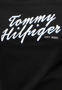 Λογότυπο Tommy Hilfiger σε λευκή γραμματοσειρά με μπλε σκιά σε μαύρο φόντο, συνοδευόμενο από το «EST. 1985» με κεφαλαία γράμματα.