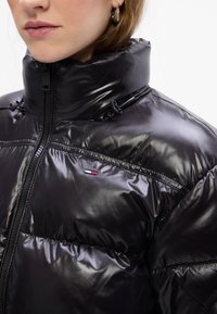 Melnā, spīdīgā pūkainā jaka ar augstu apkakli, rāvējslēdzēju un elastīgām detaļām. Iekļauj nelielu logo uzšuvumu kreisajā pusē.