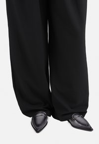 Pantofi cu vârful ascuțit din piele gri închis, cu o finisare netedă, având cusături decorative și un accent din lanț, asortați cu pantaloni negri cu croială largă.