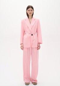 Rozā žakete un platai kāju bikses, kas izgatavotas no viegla auduma. Žaketei ir josta un viens pogas aizdare, kā arī sānu kabatas.