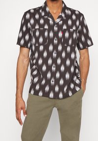 Camicia in tessuto a maniche corte di colore marrone scuro con motivo bianco a forma di piuma. Presenta un colletto, due tasche sul petto e chiusura con bottoni.