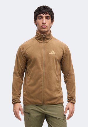 Mann trägt eine braune Fleecejacke mit Reißverschluss und Stehkragen sowie olivfarbene Cargohosen, schaut nach vorne vor einem einfarbigen Hintergrund.