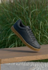 Μαύρα δερμάτινα αθλητικά παπούτσια με ομαλή υφή, με στρογγυλεμένη μύτη, μπροστινό κούμπωμα με κορδόνια και σόλα από καουτσούκ, τοποθετημένα πάνω σε ξύλινη επιφάνεια.